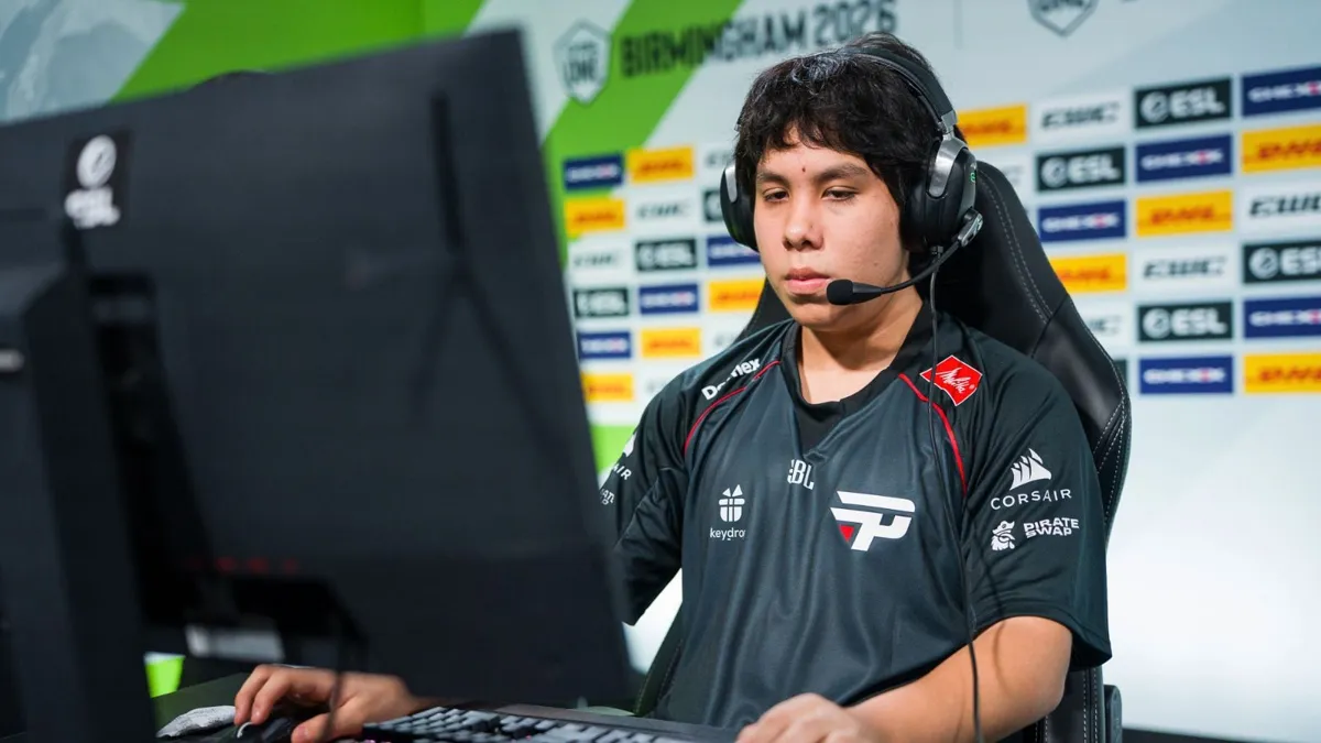 En ung man med headset sitter framför en datorskärm och spelar. Han bär en svart tröja med logotyper.